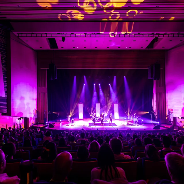 Plan large de la scène du grand auditorium du Centre de congrès éclairée dans les tons violet rose, la salle est comble.