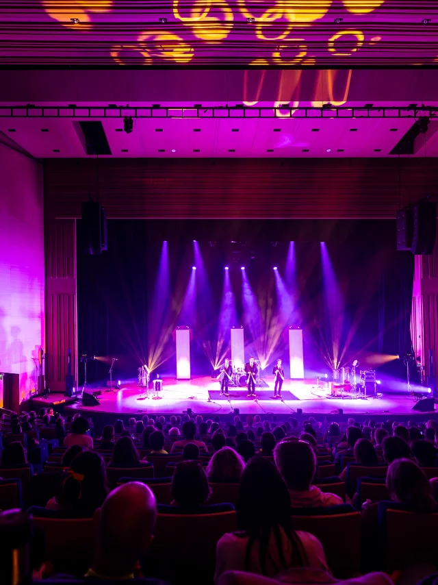 Plan large de la scène du grand auditorium du Centre de congrès éclairée dans les tons violet rose, la salle est comble.