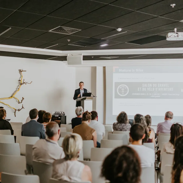 Homme au micro pupitre devant une assemblée de personnes réunies et assises