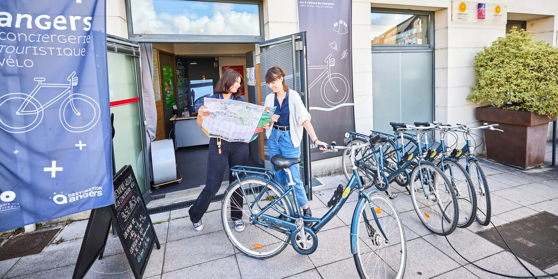 Deux jeunes femmes, l'une présente un plan à l'autre qui tient un vélo par le guidon, devant l'Office de tourisme.