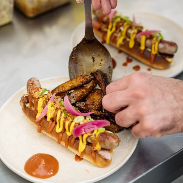 Hot dog maison préparé par le chef, gros plan sur ses mains qui dressent l'assiette.