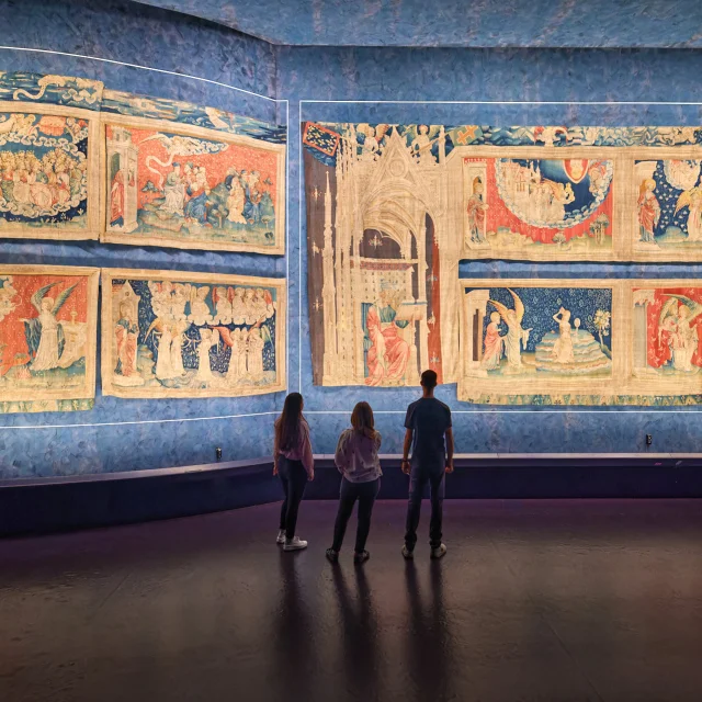 Plan large, 3 personnes de dos regardent la Tapisserie de l'Apocalypse éclairée.