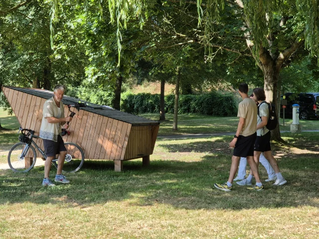 En extérieur, cameraman filmant deux jeunes hommes qui marchent dans sa direction, petite cabane en arrière plan